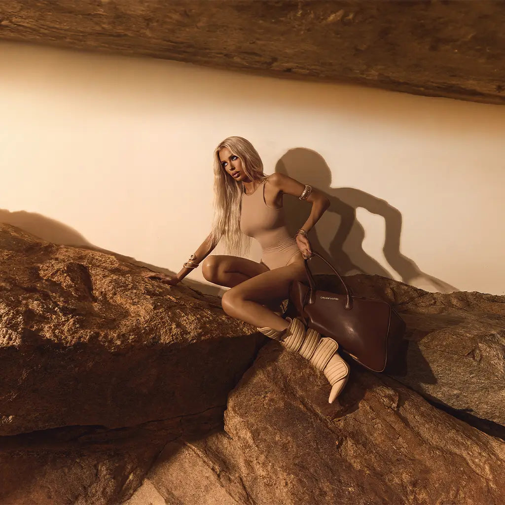 Woman sitting on a rock with a guitar, wearing a beige outfit against a neutral wall.